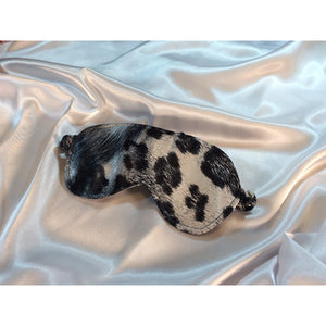 A gray, black and white satin sleep mask. The mask is lying on top of a white satin sheet.