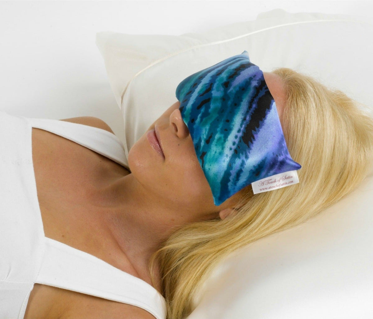 A attractive blonde haired girl lying on a white bedroom pillow with a blue pattern satin eye pillow covering her eyes.