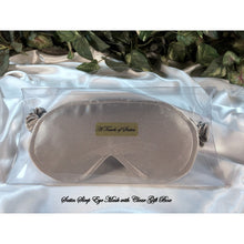 Load image into Gallery viewer, A silver satin sleep mask. The eye mask is in a clear acrylic gift box. It is placed on top of white satin sheet with various green plants in the background.