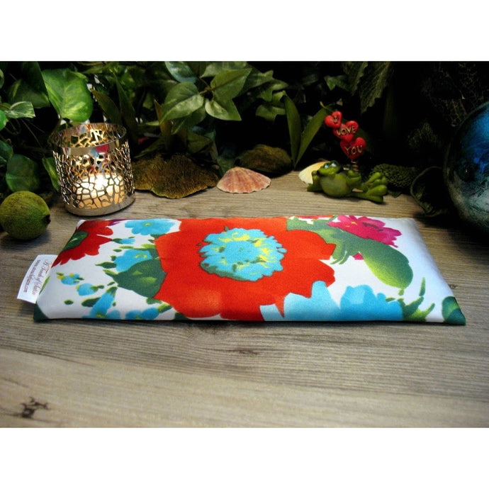 A white eye pillow with red, blue and pink flowers with splashes of green. Behind the eye pillow is a lit candle in a silver candle holder. There is also a coral colored small seashell and a small green frog holding up hearts that say I love you. In the background are various green plants.