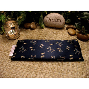 A navy blue eye pillow with a chess pieces print. Behind the eye pillow  is a lit candle in a silver candle holder. There are some smooth light colored stones and a rock with the word "Dream" etched into it. In the background there are different green leaves.
