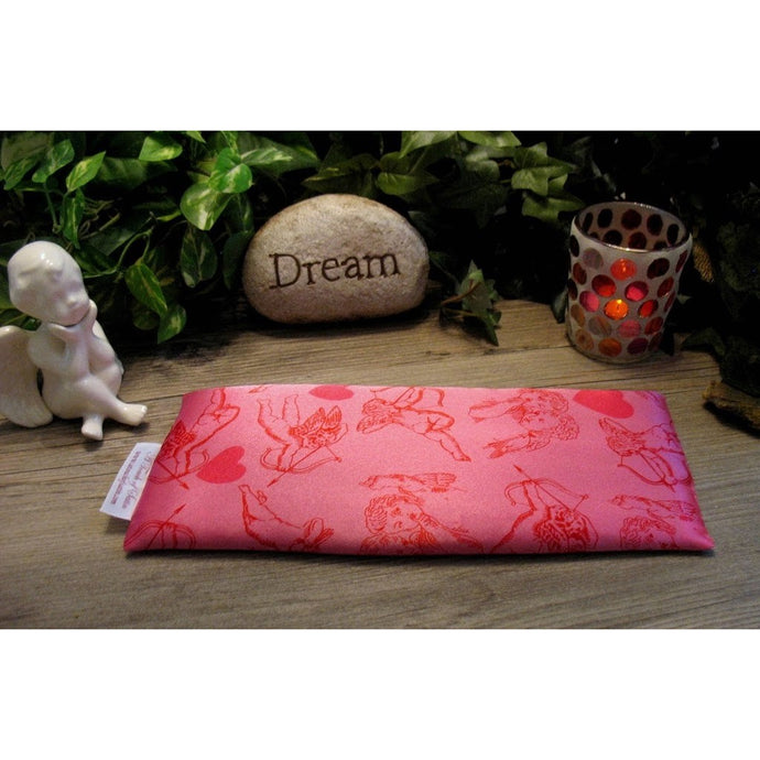 An eye pillow in pink satin with a cupid and his arrow print. Behind the eye pillow is a small white cherub statue, a beige rock that has the word 