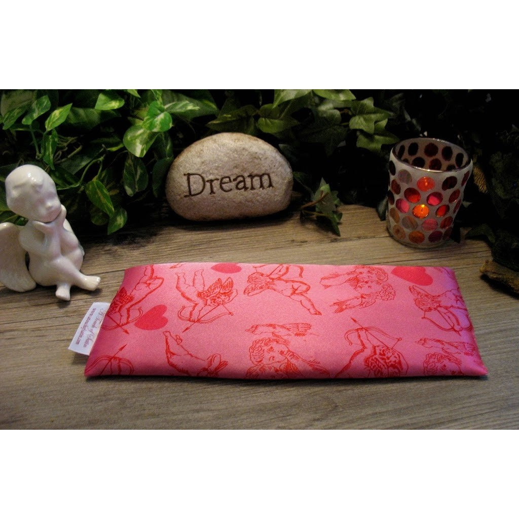 An eye pillow in pink satin with a cupid and his arrow print. Behind the eye pillow is a small white cherub statue, a beige rock that has the word 