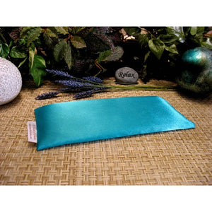 An eye pillow in turquoise blue satin. Behind it are a few sprigs of lavender and a small stone that has the word "relax" etched in it. In the background are various green leaves.
