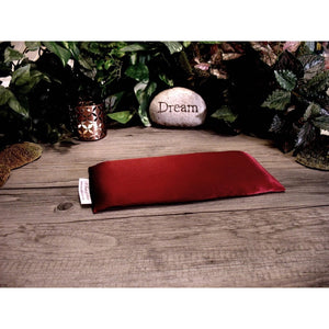 A maroon flax pillow. In the background is a candle holder, a stone with the word "dream" etched on it and various green shrubs and plants.