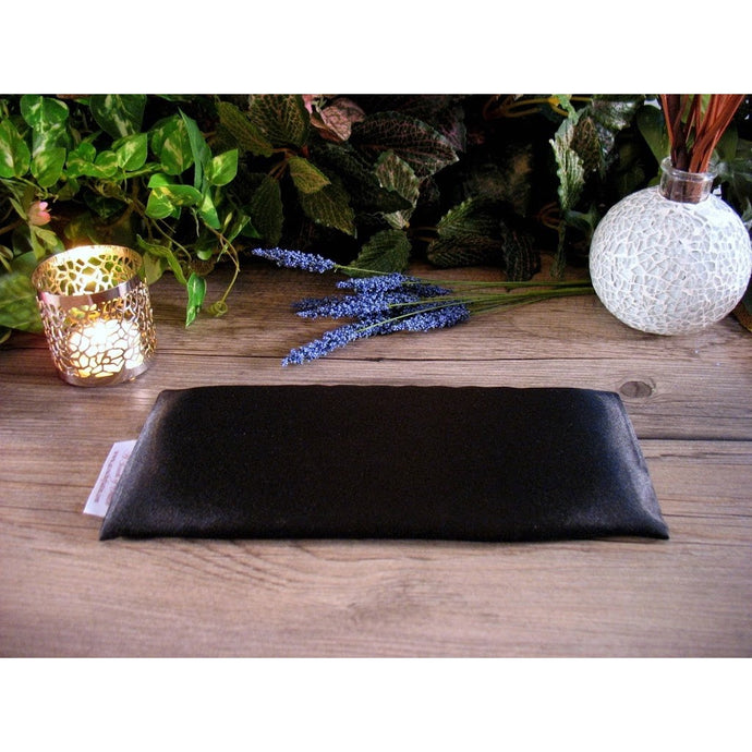 A black satin eye pillow with a candle in a candle holder burning behind it as well as some sprigs of lavender, and green foliage in the background.