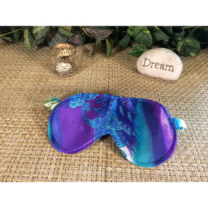 A blue and turquoise paisley print sleep mask. Behind it is a chrome candle holder and a stone with the word "dream" on it. The background has various green plants.