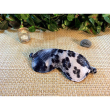 Load image into Gallery viewer, A black and gray animal print sleep mask. Behind is a chrome candle holder with a lit candle and a stone that says the word "relax" on it. In the background are various green plants.