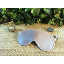 Load image into Gallery viewer, A platinum satin eye mask. Behind it is a chrome candle holder and a stone with the word &quot;relax&quot;. In the background are green plants.
