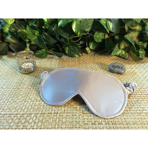 A platinum satin eye mask. Behind it is a chrome candle holder and a stone with the word "relax". In the background are green plants.