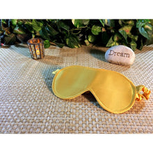 Load image into Gallery viewer, Gold sarin eye sleep mask. A candle is lit in the background and a stone with the word &quot;dream&quot; is behind the sleep mask.
