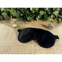 Load image into Gallery viewer, A black satin eye sleep mask with a candle burning behind it and a stone with the word "relax" in back of it.