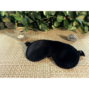 A black satin eye sleep mask with a candle burning behind it and a stone with the word "relax" in back of it.
