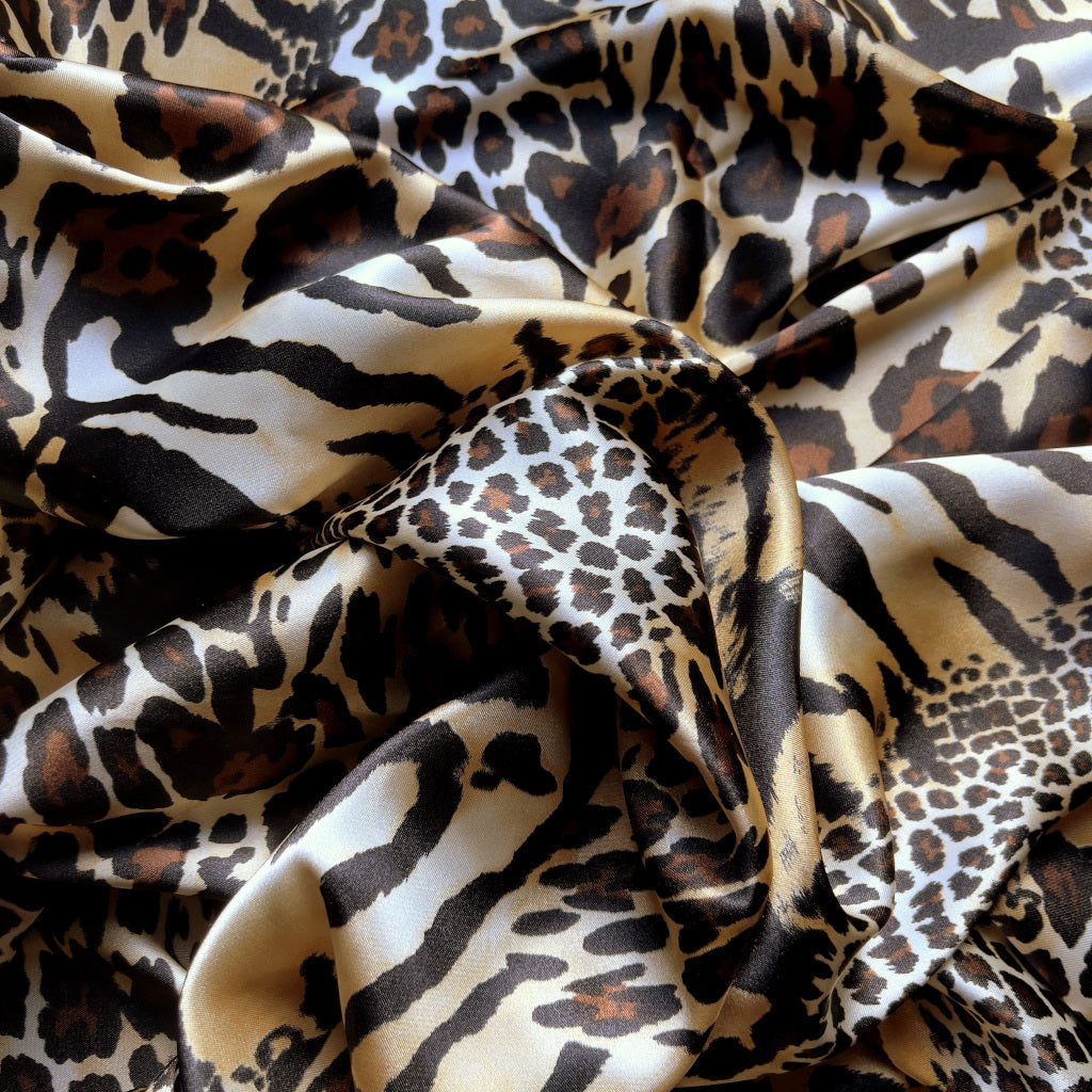 A view from above looking down at a brown, beige, black and gold cheetah and leopard print satin fabric. Twisted in a swirl to show the pattern.