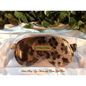 A brown and gold leopard print satin sleep mask. The eye mask is in a clear acrylic gift box. It is placed on top of white satin with various green plants in the background.