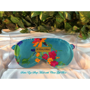 A turquoise satin sleep mask with yellow and pink flowers. The sleep mask is in a clear acrylic gift box. It is placed on top of white satin with various green plants in the background.