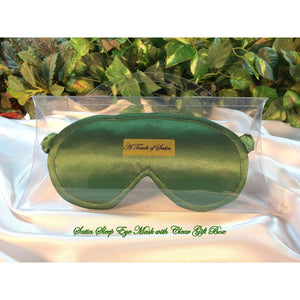 A sage green satin sleep mask. The sleep mask is in a clear acrylic gift box. There is an A Touch of Satin label on the front of the gift box. It is placed on top of white satin sheets with various green plants in the background.