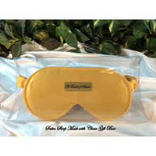 Load image into Gallery viewer, A gold satin sleep mask. The sleep mask is in a clear acrylic gift box. It is placed on top of white satin with various green plants in the background.
