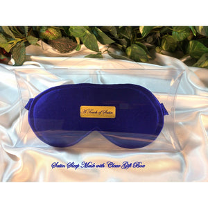A solid blue satin sleep mask. The sleep mask is in a clear acrylic gift box. There is an A Touch of Satin label on the front of the gift box. It is placed on top of white satin with various green plants in the background.