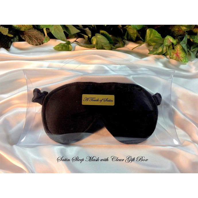 A black satin sleep mask. The sleep mask is in a clear acrylic gift box. It is placed on top of white satin sheet with various green plants in the background. A gold label that says A Touch of Satin is in the middle of the gift box and belowthe gift box are the words 