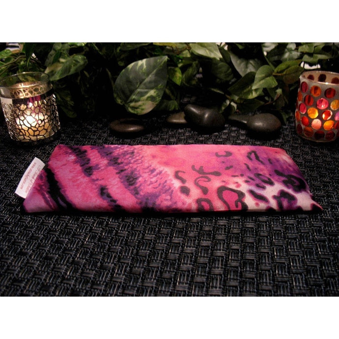 A pink, African animal print eye pillow. Behind it is a lit candle on either side in two different candle holders. There are some dark bron smooth stones between the candle holders. In the background are different green plants. 