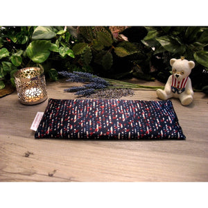 A red, white and blue print eye pillow. Behind the eye pillow is a lit candle in a silver candle holder. There are some sprigs of lavender and to the right of that is a small ceramic white bear wearing the stars and stripes. In the background is a variety of green plants.
