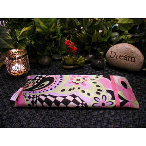 An eye pillow in pink, light green, white and black. It has a hippie, Boho, Mod vibe or feel. Behind the eye pillow is a lit candle in a silver candle holder. Next to that is a frog holding small hearts that say "I love you". Next to the frog is a light colored stone with the word "Dream" etched into it. In the background are various green plants.