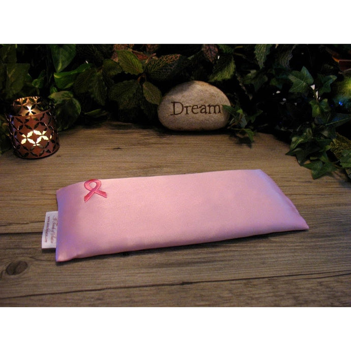 An eye pillow in solid pink with the ribbon logo for breast cancer in the top corner of the eye pillow. Behind the eye pillow is a lit candle in a bronze candle holder and on the other side is a light colored stone with the word 