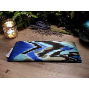 An eye pillow in a blue, black and gray southwestern print. Behind the eye pillow is a lit candle in a silver candle holder. There are also some sprigs of lavender. In the background are different shades of green leaves.