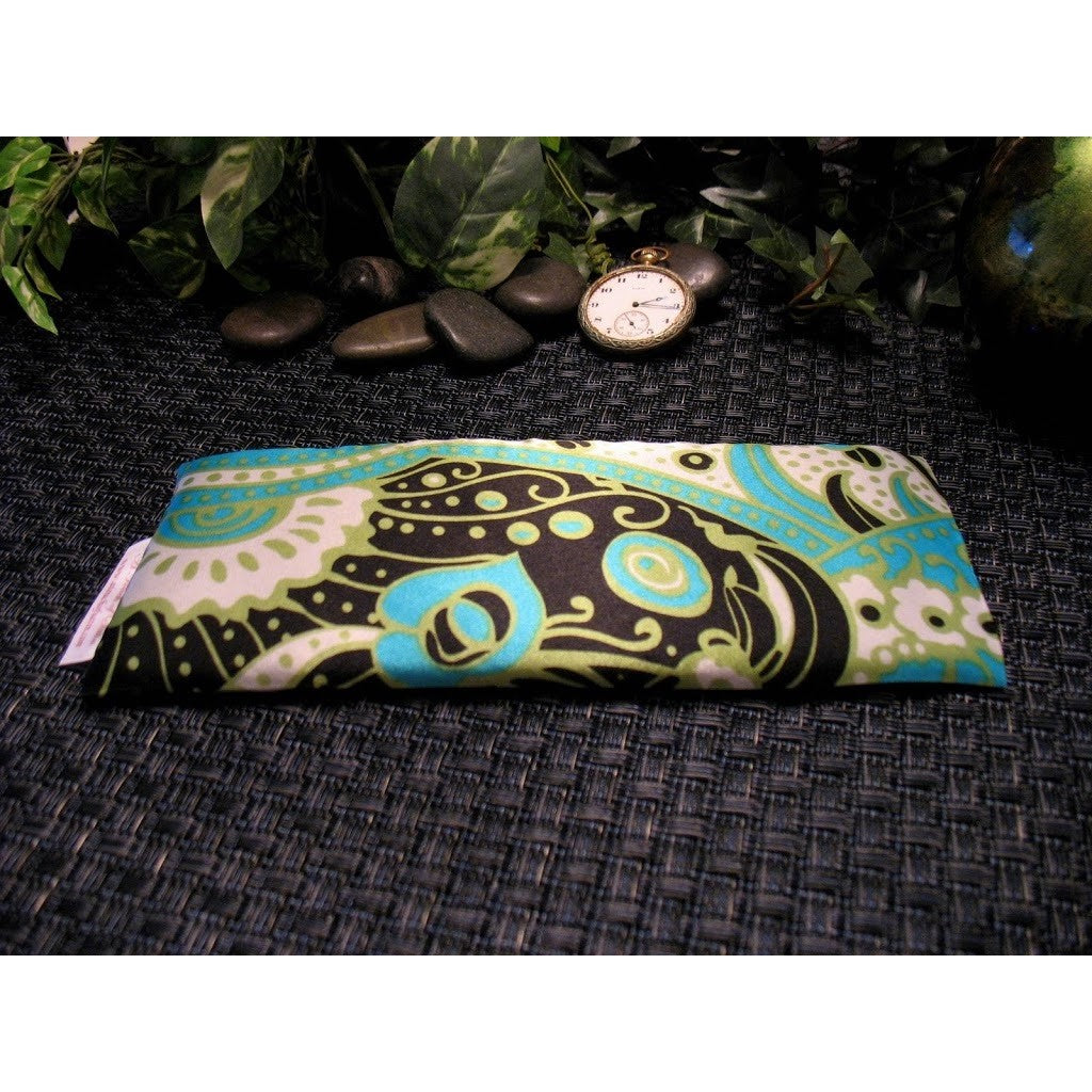 An eye pillow with a green, white, black and blue abstract print. Behind the eye pillow are a few smooth dark rocks with a pocket watch resting on them. The time on the watch is 2:15.  In the background are various green leaves.