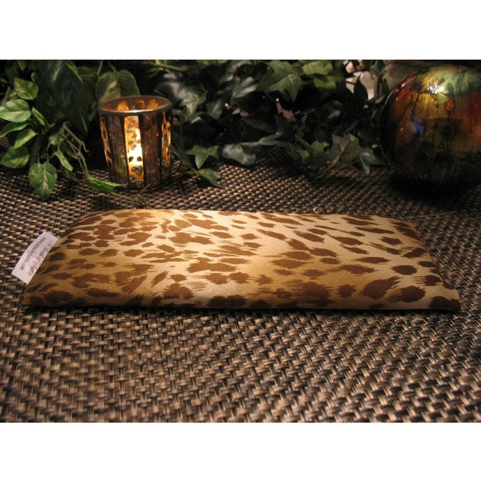 An eye pillow with a brown and gold cheetah animal print. Behind the eye pillow is a lit candle in a gold and brown glass candle holder. A brown, green and gold ceramic ball is on the other side from the candle holder. In the background are various green plants.