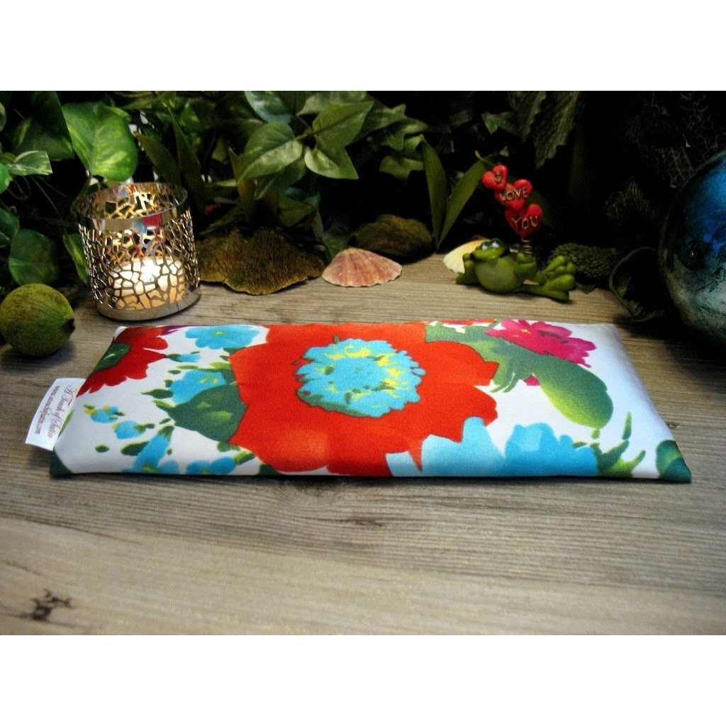 A white eye pillow with red, blue and pink flowers with splashes of green. Behind the eye pillow is a lit candle in a silver candle holder. There is also a coral colored small seashell and a small green frog holding up hearts that say I love you. In the background are various green plants.