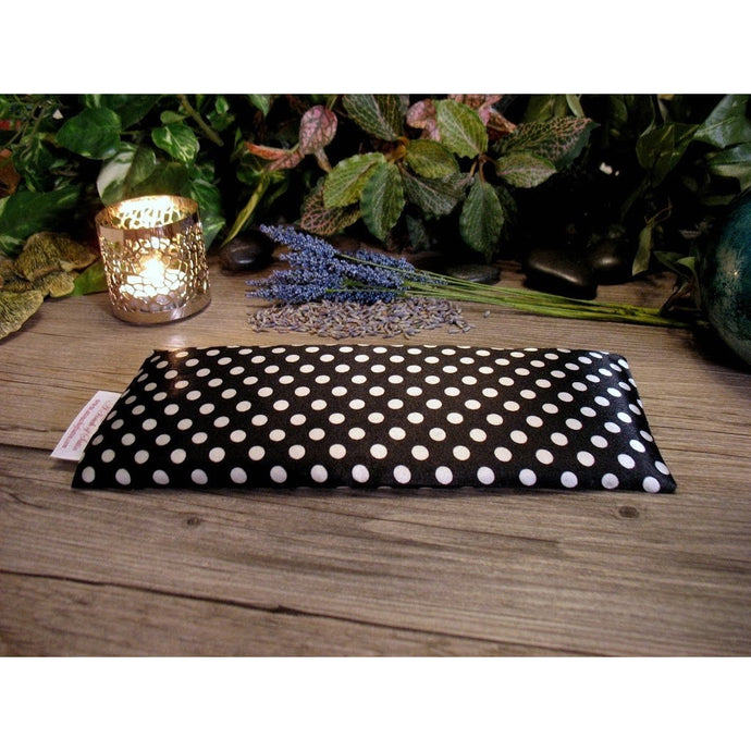 A black with white polka dots eye pillow. Behind the eye pillow is a lit candle in a silver candle holder. There are sprigs of lavender and lavender buds. In the background are various green and pink leaves.