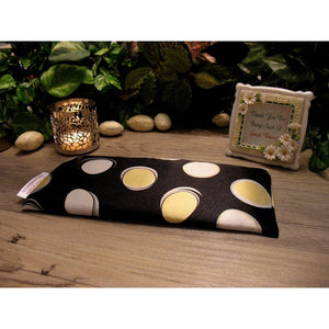 An eye pillow in black satin with large yellow and white polka dots. There is a lit candle in a silver candle holder and a framed picture with a saying about friends behind the eye pillow. Various green plants are in the background.