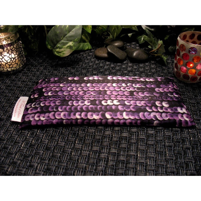 An eye pillow with a purple and black sequin satin print, Behind the eye pillow are two lit candles in candle holders. One on each side. In the background are a couple of smooth dark stones and various green leaves.
