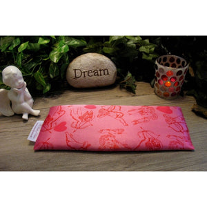 An eye pillow in pink satin with a cupid and his arrow print. Behind the eye pillow is a small white cherub statue, a beige rock that has the word "Dream" etched into it and a lit candle in a candle holder. Green plants are in the background.