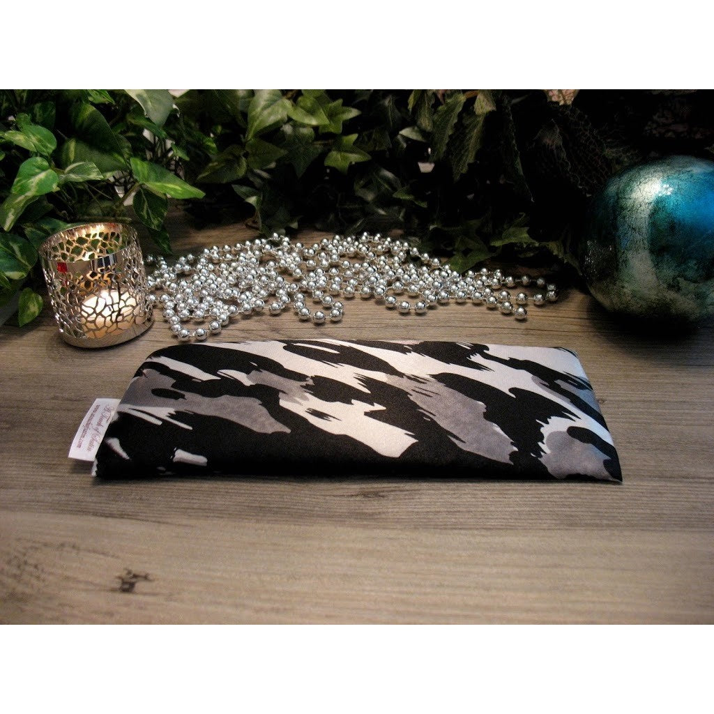 A black, gray and white print eye pillow. There is a lit candle in a silver candle holder, and white pearl neckless and various green plants in the background.