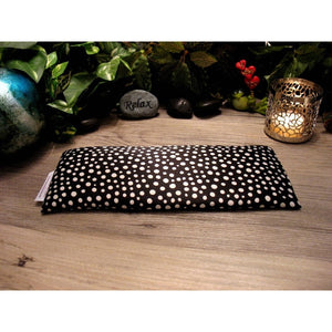 A black eye pillow with various small white dots. Behind the eye pillow is a small gray stone that has the word "relax" etched on it and a lit candle in a silver candle holder. There is a small green frog holding a sign that says "I love you".  Green foliage is in the background.