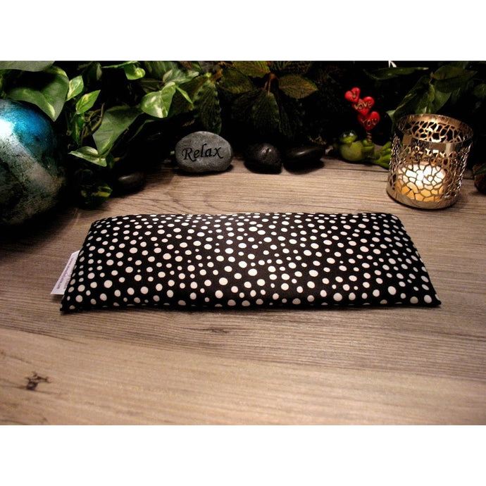 A black eye pillow with various small white dots. Behind the eye pillow is a small gray stone that has the word 