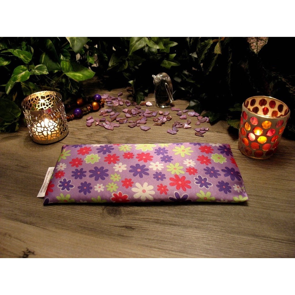 An eye pillow with purple satin material that has white, pink, green and dark purple daisies. Behind are two lit candles in candle holders. In the background various green plants.
