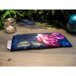 A navy blue eye pillow with a pink rose and green leaves. Behind the eye pillow is a lit candle in a silver candle holder. There are a few sprigs of lavender and some lavender buds. In the background are various green plants. 