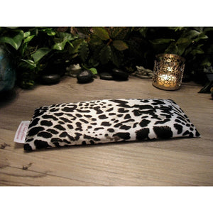 A black and white leopard print eye pillow. Behind it is a lit candle in a silver candle holder. In the background is various green plants.