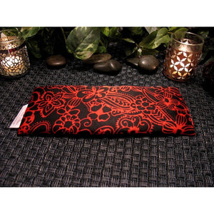 A black with red flowers eye pillow. Behind are two lit candles in a bronze candle holder and a silver one. There are a few polished, dark stones. A variety of green leaves are in the background.