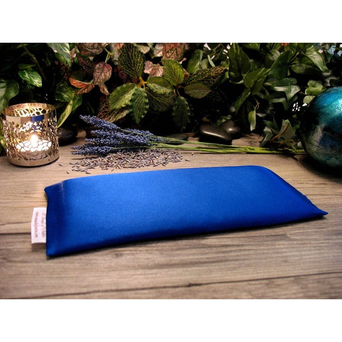A royal blue eye pillow. Behind it is a lit candle in a silver candle holder. There are some sprigs of lavender and some loose lavender buds. In the background are various green plants.