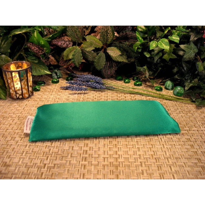 An eye pillow in a Kelly green color. Behind it is a lit candle in a gold and brown candle holder and a few sprigs of lavender. In the background are a few green gemstones and a variety of different green leaves.