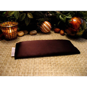 A chocolate brown eye pillow. Behind it is a burning candle in a gold and bronze candle holder, a few light colored rocks and a couple of tropical nuts. There are various green leaves in the background.