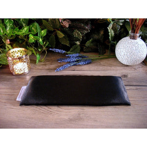 A black satin eye pillow with a candle in a candle holder burning behind it as well as some sprigs of lavender, and green foliage in the background.