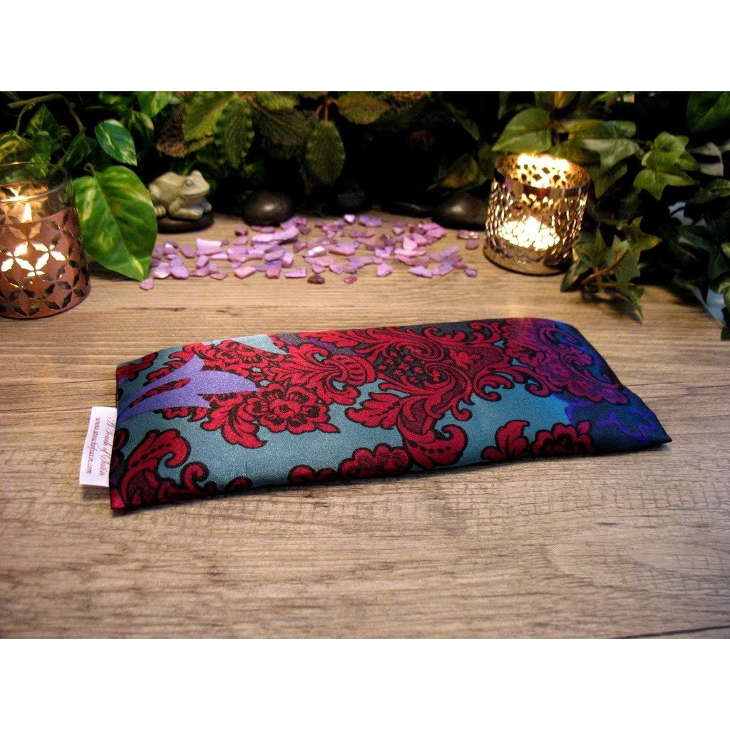 An eye pillow with a red and green paisley print that has a hint of purple in it. Behind it are two lit candles in candle holders. There are some very small light purple  stones. In the background are different shades of green leaves.