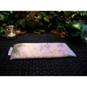 An eye pillow with pink and lavender flowers and soft, green leaves. Behind the eye pillow is a lit candle in a silver candle holder. To the right is a small green and blue ceramic ball. In the background are various green plants.