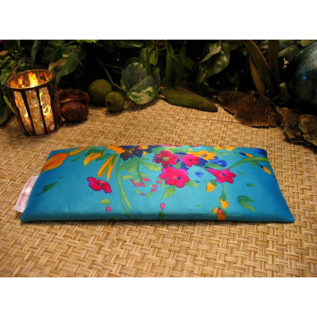 An eye pillow with a turquoise blue with pink flowers print. Behind the eye pillow is a lit candle in a candle holder. Green plants are in the background.